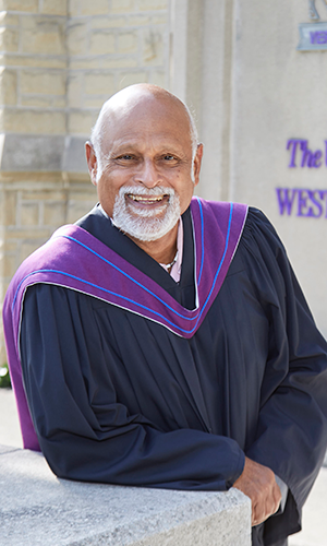 Photo of Anton Allahar leaning on a brick wall wearing his academic robes.