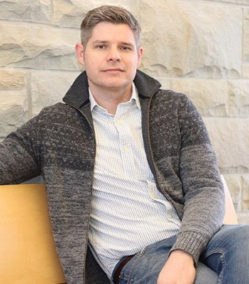 Photo of Warren Steele wearing a grey cardigan and sitting on a yellow bench.