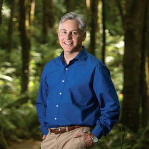 John Borrows wearing a blue shirt standing in a forest surrounded by trees
