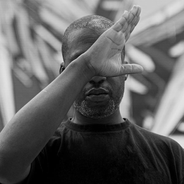 black and white photo of a black man with short hair wearing a black t-shirt. His arm bent upward in front of face with his fingers outstretched. 
