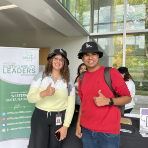 two students standing in front of sustainability banner with sustainability bucket hats